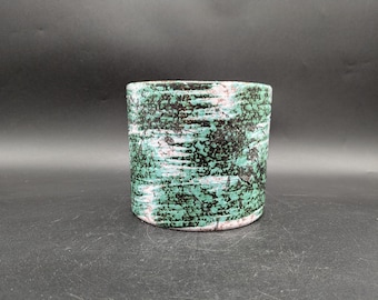 Puede incluir: Una maceta cilíndrica de cerámica con superficie texturizada. La maceta presenta un patrón moteado de color turquesa, negro y blanco, creando una estética orgánica única. Adecuada para plantas o como objeto decorativo.