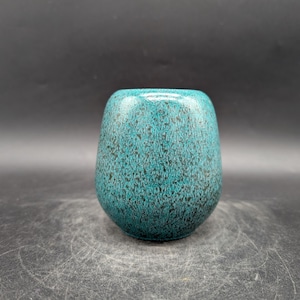 Könnte beinhalten: Eine türkisfarbene Keramikvase mit einem gesprenkelten Muster. Die Vase hat eine abgerundete, leicht konische Form und eine glänzende Oberfläche. Das gesprenkelte Design weist dunkle Flecken im gesamten türkisfarbenen Glanz auf.