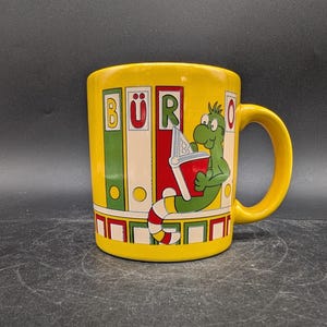 May include: Yellow ceramic mug with a green cartoon dragon reading a book. The mug has the word "BÜR" printed on it, along with colorful book-like designs. The handle and interior are also yellow.