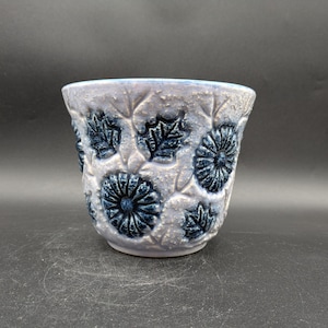 May include: A light purple ceramic planter with a textured surface and a floral design in dark blue. The planter features raised flowers and leaves.
