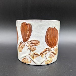 May include: White ceramic planter with brown floral design. The planter has a cylindrical shape and features a raised design of stylized flowers.
