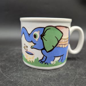 Könnte beinhalten: Weiße Keramik-Tasse mit einem blauen Cartoon-Elefanten mit grünem Ohr und einer rosa-gelben Decke. Der Elefant steht auf grünem Gras. Die Tasse hat einen weißen Henkel und einen weißen Innenraum.