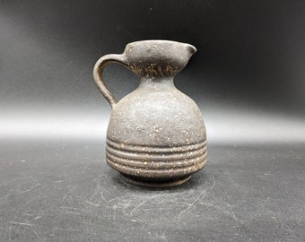 May include: A small, dark brown ceramic pitcher with a speckled glaze and a rounded body. The pitcher has a handle and a spout.