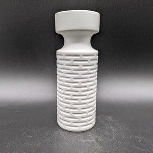 Könnte beinhalten: Eine weiße Keramikvase mit einem strukturierten, geometrischen Muster. Die Vase hat einen breiten, runden Boden und einen schmalen Hals.