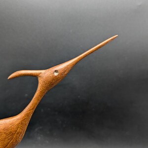 May include: A wooden bird sculpture with a long, slender beak and a curved neck. The bird is a warm brown color with a small, round, inlaid detail near the eye. The sculpture is set against a dark background.