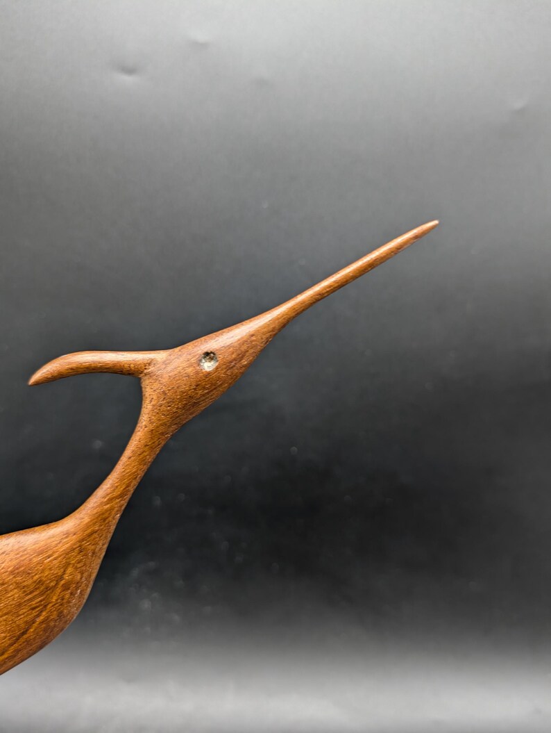 May include: A wooden bird sculpture with a long, slender beak and a curved neck. The bird is a warm brown color with a small, round, inlaid detail near the eye. The sculpture is set against a dark background.