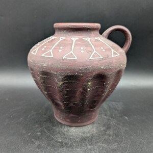May include: A maroon ceramic pitcher with a small handle and a rounded body. The pitcher features a white geometric design around the neck and a textured surface. The base has a ribbed design.