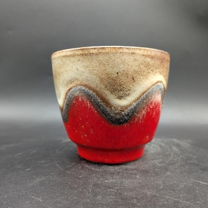Könnte beinhalten: Eine kleine Keramik-Tasse mit einem einzigartigen Design. Der obere Teil ist hellbraun, geht in ein wellenförmiges schwarz-weißes Band über und hat einen leuchtend roten Boden. Die Tasse hat einen leicht ausgestellten Rand.