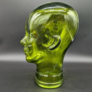 Puede incluir: Una cabeza de maniquí de vidrio verde con una superficie lisa y redondeada. La cabeza está ligeramente inclinada hacia un lado y los ojos están cerrados. La cabeza está sentada sobre una superficie negra.