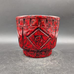 Könnte beinhalten: Ein rot-schwarzer Keramik-Pflanztopf mit geometrischen Mustern. Der Pflanztopf hat eine rautenförmige Form mit einem Kreis in der Mitte. Die Außenseite hat ein strukturiertes Aussehen. Geeignet für den Innen- und Außenbereich.