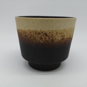 Puede incluir: Un macetero de cerámica marrón y beige con un esmalte moteado. El macetero tiene una abertura redonda ancha y una base ligeramente acampanada.