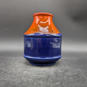 Può includere: Un vaso di ceramica con coperchio. Il vaso è blu scuro con la parte superiore arancione. Il vaso ha una forma arrotondata con la parte superiore leggermente svasata. La superficie ha una finitura lucida.