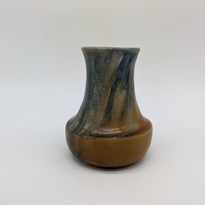 Könnte beinhalten: Eine kleine, braune und blaue Keramikvase mit einer ausgestellten Öffnung. Die Vase hat eine gefleckte Glasur mit blauen und braunen Streifen.
