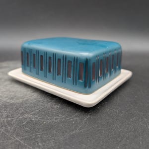 Könnte beinhalten: Eine türkisfarbene Keramik-Butterdose mit einem rechteckigen Deckel mit vertikalen schwarzen Streifen. Die Dose steht auf einer weißen, quadratischen Basis. Es ist ein Küchenartikel.