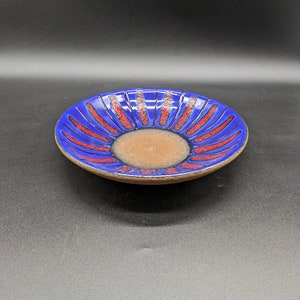 Puede incluir: Un plato de cerámica con un exterior azul vibrante y un centro marrón. Patrones lineales rojos irradian desde el centro, creando un diseño de estallido solar. El plato tiene un borde ligeramente elevado y una apariencia rústica hecha a mano.