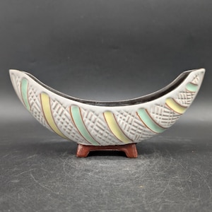 May include: A ceramic, boat-shaped bowl with a gray exterior, accented with yellow and green stripes. The bowl has a woven texture and sits on a small, brown base. The interior is black.