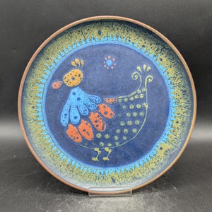May include: A round ceramic plate with a dark blue center featuring a stylized bird design in blue, orange, and yellow. The rim has a textured pattern in green and yellow, with a brown edge. The plate is on a clear stand.