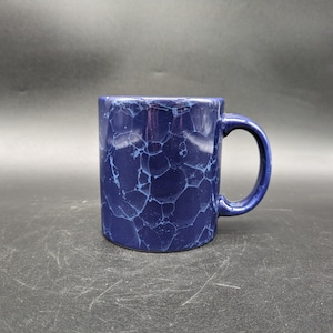 Puede incluir: Una taza de cerámica azul con un patrón de esmalte craquelado. La taza tiene una asa redondeada.