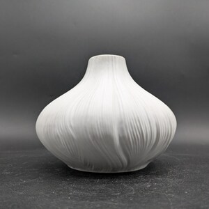 Könnte beinhalten: Eine weiße Keramikvase mit einem strukturierten, gerippten Design. Die Vase hat eine abgerundete, bauchige Form.