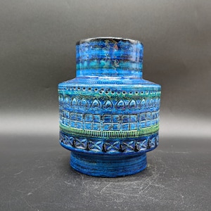 Op de afbeelding: Een blauwe keramische vaas met een textuur en een cilindrische vorm. De vaas heeft geometrische patronen in blauw, groen en zwart. De vaas is ongeveer 20 cm hoog.