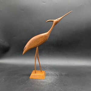 May include: A wooden sculpture of a bird with a long neck and beak, standing on a square base. The bird is a warm brown color, with a smooth, polished finish. The sculpture is a decorative item.