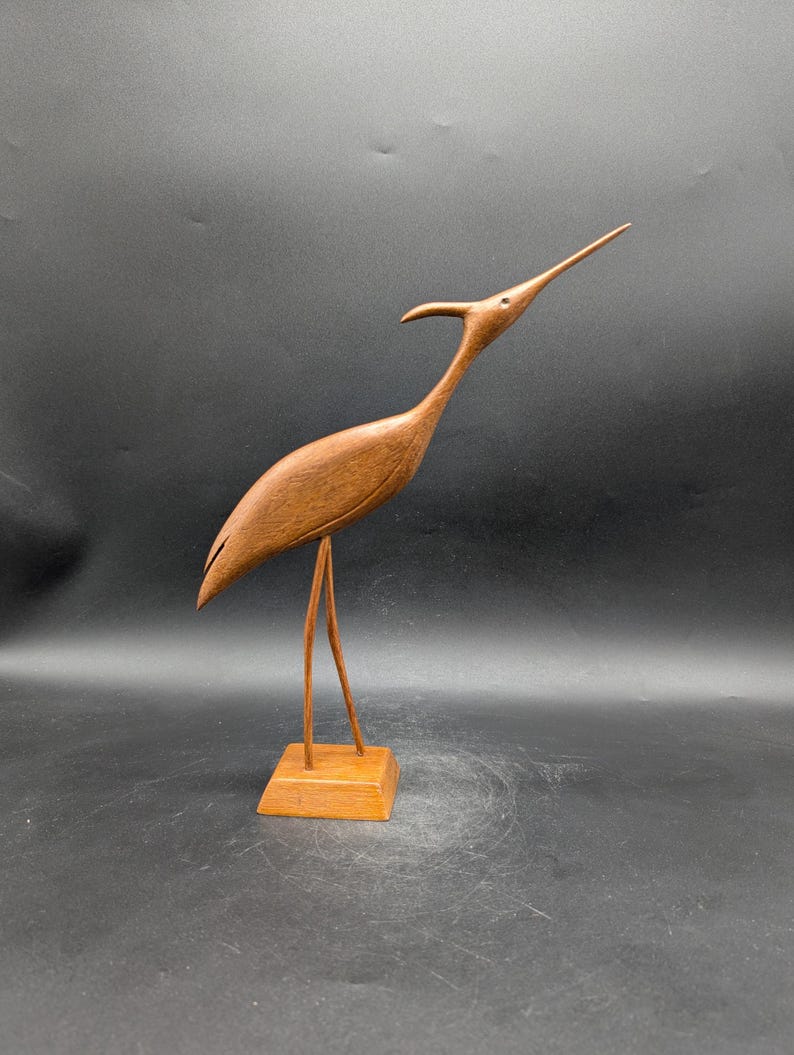 May include: A wooden sculpture of a bird with a long neck and beak, standing on a square base. The bird is a warm brown color, with a smooth, polished finish. The sculpture is a decorative item.