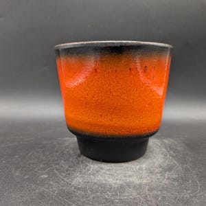 Op de afbeelding: Een keramische beker met een opvallende kleurovergang. De bovenste rand is zwart, gaat over in een feloranje lichaam en eindigt met een zwarte basis. De beker heeft een getextureerd oppervlak.