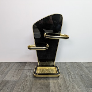 Puede incluir: Un soporte de baño negro y dorado con tres estantes. El soporte tiene un diseño geométrico y una base dorada.