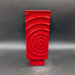 Könnte beinhalten: Eine rechteckige, rote Keramikvase mit strukturierter Oberfläche. Die Vase hat ein Spiralmuster auf der Vorderseite. Der Sockel ist ein roter Block. Die Vase eignet sich zur Präsentation von Blumen oder als dekoratives Element. Die Vase ist etwa 20 cm hoch.