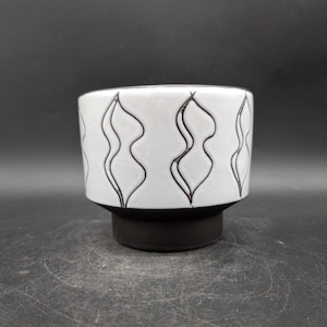 Könnte beinhalten: Weißer Keramik-Pflanztopf mit schwarzem Sockel und sich wiederholendem schwarzem Wellenlinien-Design. Der Pflanztopf hat eine zylindrische Form und eignet sich für kleine Pflanzen oder Sukkulenten.