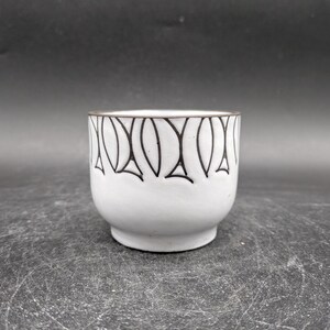 Könnte beinhalten: Weiße Keramiktasse mit einem schwarzen geometrischen Muster am Rand.