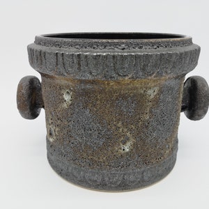 May include: A dark grey ceramic planter with two rounded handles on the sides. The planter has a textured surface and a rough, uneven finish.