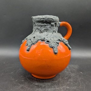 Puede incluir: Una jarra de cerámica con un cuerpo naranja vibrante y un borde y asa gris oscuro texturizados. El material gris parece estar goteando sobre la superficie naranja. La jarra tiene una forma redondeada y un asa pequeña.