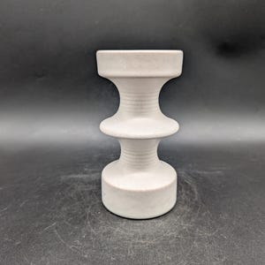 May include: A light grey ceramic candle holder with an hourglass shape. The candle holder has a wide base, a narrow middle section, and a wide top. The surface has a textured, matte finish. The design is minimalist and modern, suitable for various decor styles.