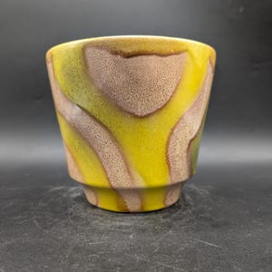 May include: A ceramic planter with a unique design. The exterior features a blend of yellow, brown, and green hues in a wavy pattern. The planter has a cylindrical shape with a slightly flared base, suitable for small plants or decorative use.