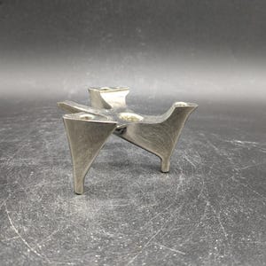May include: A silver metal candle holder with a unique, abstract design. The holder has three legs and a central point for holding a candle.