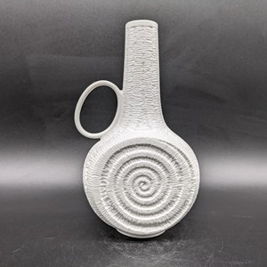 Könnte beinhalten: Weiße Keramikvase mit strukturierter Oberfläche und Spiralmuster. Die Vase hat einen Henkel und einen runden Korpus.