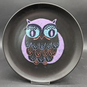 Könnte beinhalten: Schwarzer Keramikteller mit einem violetten Eulenmotiv. Die Eule hat blaue Augen, schwarze Federn und einen rosa Schnabel.