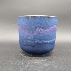 Peut inclure: Une tasse en céramique faite à la main avec une surface texturée. La tasse présente un dégradé de nuances de bleu, avec une bande violette au milieu. Le bord est d'une couleur plus foncée, presque noire. La tasse est une pièce de poterie unique.