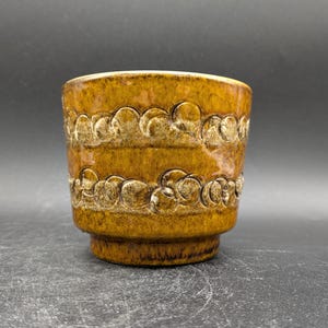 Könnte beinhalten: Ein brauner Keramiktopf mit einem strukturierten Design aus erhabenen Kreisen. Der Topf hat einen leicht ausgestellten Rand und einen abgerundeten Boden.