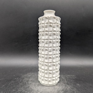 Puede incluir: Un jarrón alto de cerámica blanca con un patrón geométrico texturizado. El jarrón tiene un cuerpo cilíndrico y un cuello estrecho. La superficie está cubierta con un patrón repetitivo de formas tridimensionales en relieve. Es un objeto decorativo.