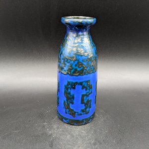 Puede incluir: Un pequeño jarrón de cerámica azul con un esmalte moteado negro y azul. El jarrón tiene un patrón geométrico azul en la parte delantera.