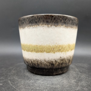 Könnte beinhalten: Eine Keramiktasse mit einem geschichteten Design. Die oberen und unteren Abschnitte sind dunkelbraun, mit einem weißen und olivgrünen Band in der Mitte. Die Tasse hat eine zylindrische Form und einen leicht ausgestellten Boden.