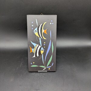 Könnte beinhalten: Eine schwarze Keramikfliese mit einem bemalten Design aus zwei schwarz-weißen Fischen mit blauen und gelben Akzenten, die vor grünen und blauen Algen schwimmen. Die Fliese hat oben eine Aufhängeschlaufe.