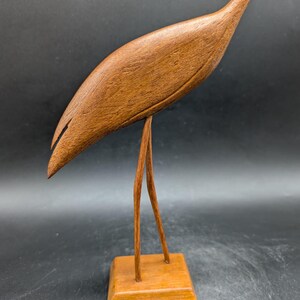 May include: A wooden sculpture of a bird with a long neck and beak. The bird is brown and stands on two thin legs attached to a square base. The sculpture is made of wood and has a smooth finish. The bird's body is curved, and its head is tilted downward.