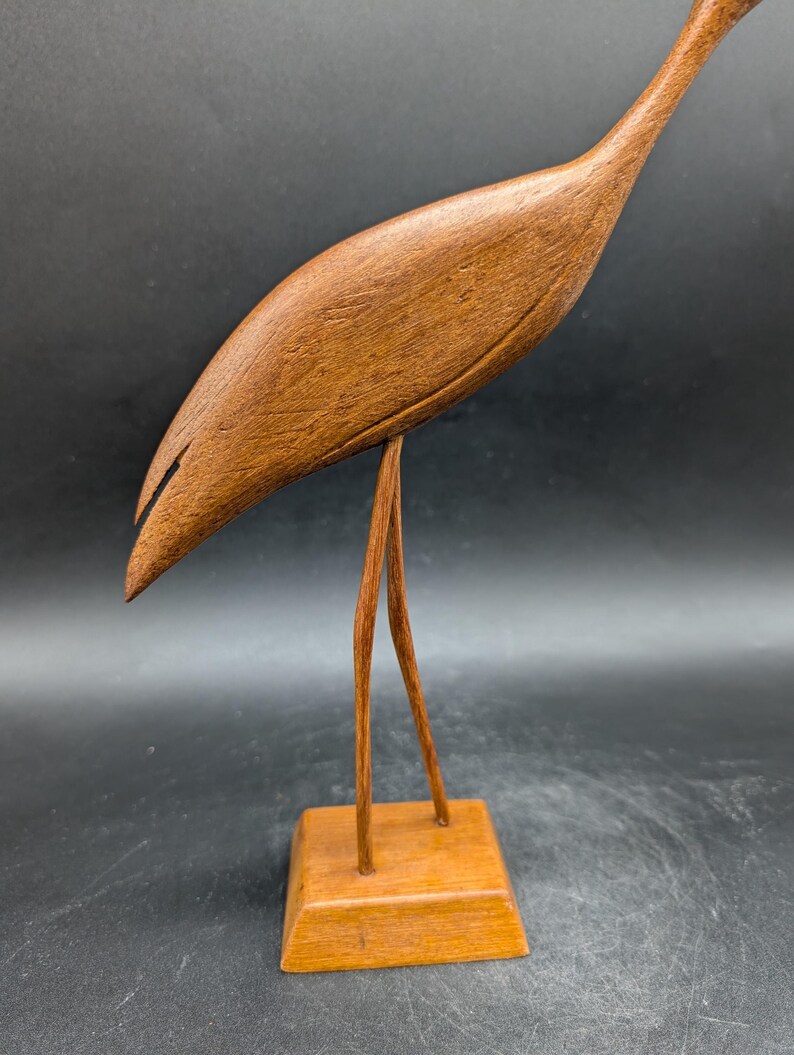 May include: A wooden sculpture of a bird with a long neck and beak. The bird is brown and stands on two thin legs attached to a square base. The sculpture is made of wood and has a smooth finish. The bird's body is curved, and its head is tilted downward.