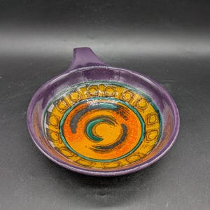 Puede incluir: Un plato de cerámica morado con asa. El interior presenta un diseño vibrante con patrones en espiral naranja, amarillo y verde. El plato tiene un acabado brillante y probablemente está hecho a mano.