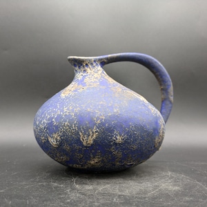 May include: A blue ceramic pitcher with a rounded body and a curved handle. The pitcher has a textured surface with a speckled, gold-colored pattern. The rim of the pitcher is also gold-colored.
