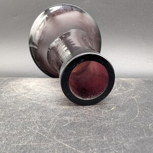 May include: A dark purple glass vase with a rounded body and a narrow neck. The vase has a black rim around the opening. The interior of the vase is a deep red color. The vase is lying on a textured surface.