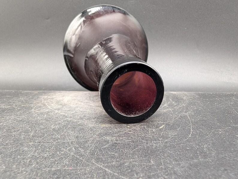 May include: A dark purple glass vase with a rounded body and a narrow neck. The vase has a black rim around the opening. The interior of the vase is a deep red color. The vase is lying on a textured surface.